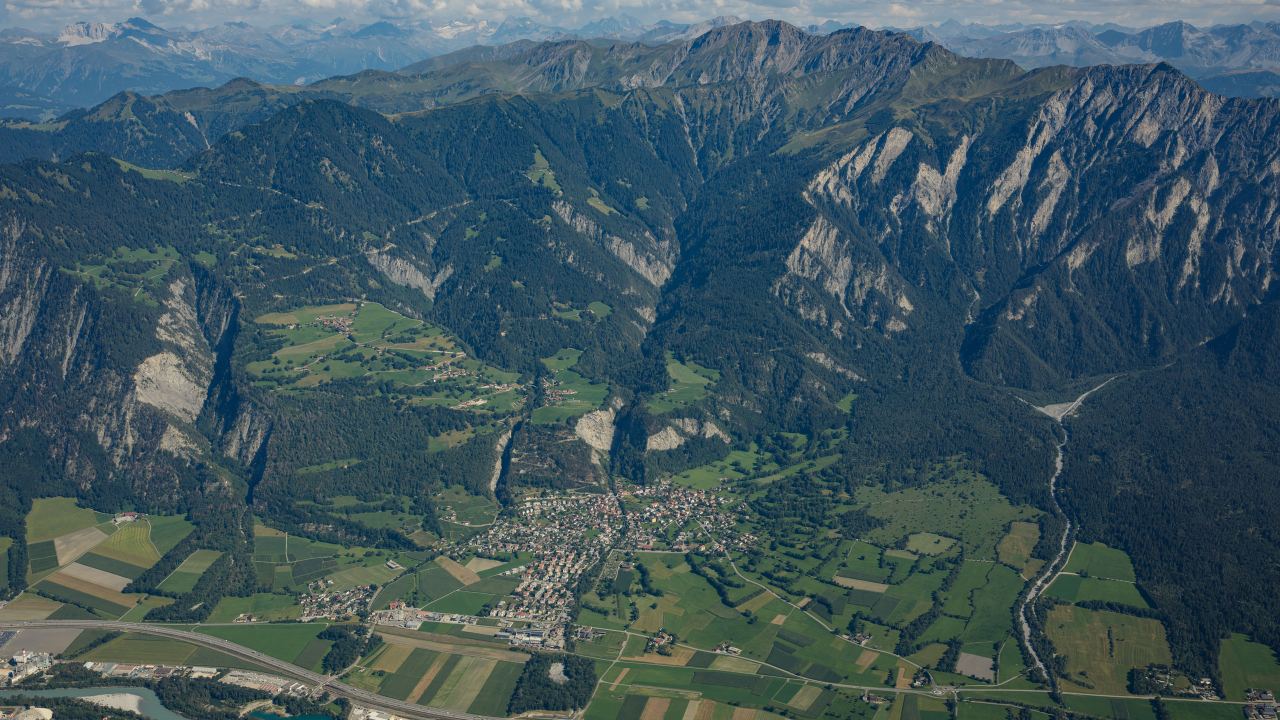 Schrägluftbild des Rheintals bei Trimmis, steile Bergflanken mit Rüfen über Dorf, Feldern und Autobahn