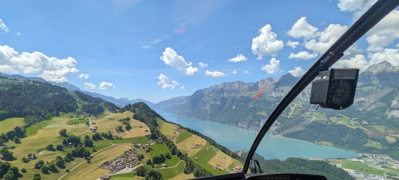 Blick aus dem Helikoptercockpit über einen Bergsee, rechts der Kompass am Scheibenrahmen