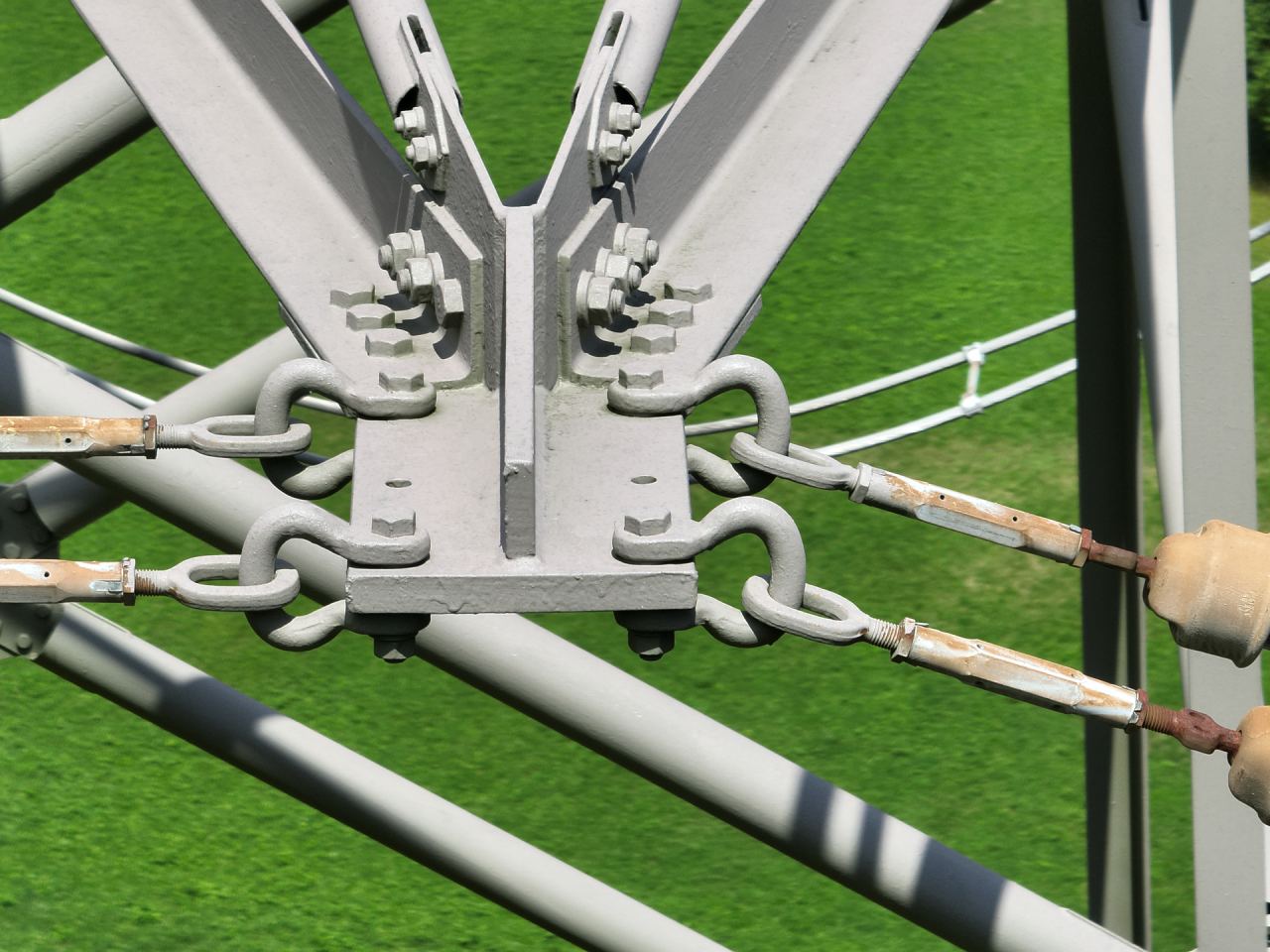 Detailaufnahme der Aufhängung von Isolatorenketten an einem Freileitungsmast, mit verschraubtem Knotenblech, Schäkeln und Gewindestangen vor einer Wiese